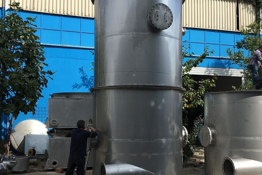 Mavi binanın önünde, açık alanda dik duran büyük paslanmaz tank; dibinde çalışan usta